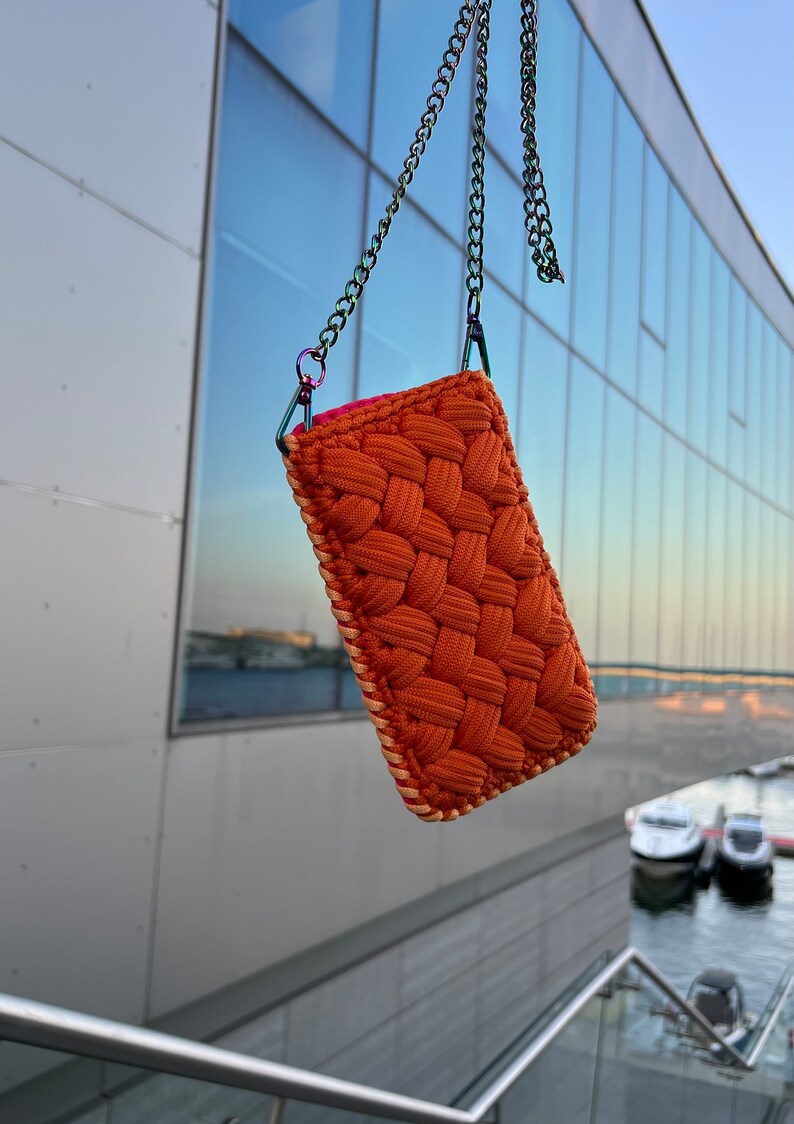 May include: A rectangular pouch crafted from thick, woven orange cord with a textured, braided pattern. The pouch features a light-colored stitched border and is suspended by a metallic, iridescent chain strap attached with matching hardware.