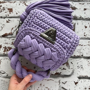 Puede incluir: Un bolso de ganchillo color lavanda con un asa gruesa trenzada y una borla grande a juego. El bolso est&aacute; confeccionado con hilo de tela con un patr&oacute;n de punto texturizado e incluye un cierre de giro de metal plateado en la solapa frontal.