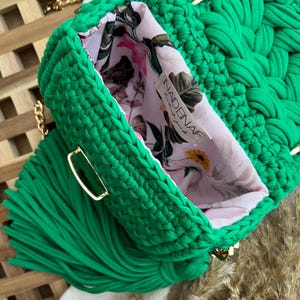 Puede incluir: Un bolso de ganchillo verde brillante con una textura trenzada gruesa, una correa de cadena dorada y una gran borla verde a juego. El interior est&aacute; forrado con una tela rosa claro estampada con un motivo floral y una etiqueta que dice NADENAF.