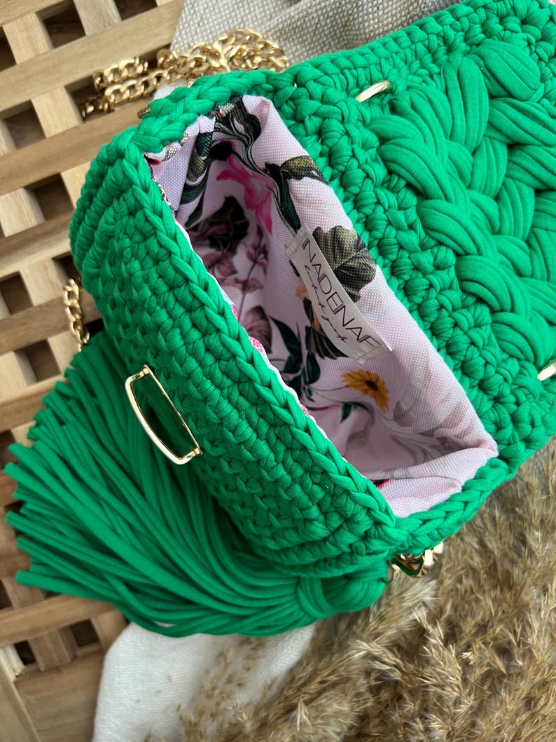 Puede incluir: Un bolso de ganchillo verde brillante con una textura trenzada gruesa, una correa de cadena dorada y una gran borla verde a juego. El interior est&aacute; forrado con una tela rosa claro estampada con un motivo floral y una etiqueta que dice NADENAF.