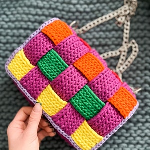 May include: A rectangular crochet handbag featuring a woven pattern of vibrant yellow, orange, green, and magenta yarn squares. The bag is bordered with light purple yarn and includes a silver-toned metal chain strap attached to the top.