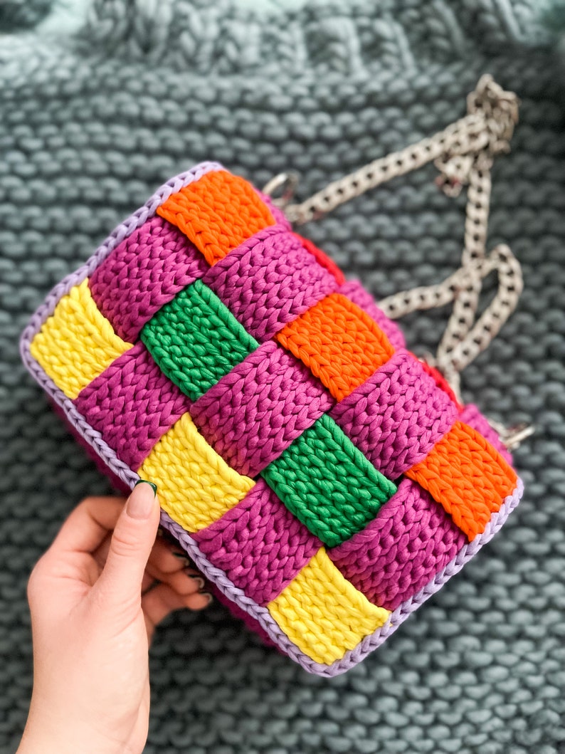 May include: A rectangular crochet handbag featuring a woven pattern of vibrant yellow, orange, green, and magenta yarn squares. The bag is bordered with light purple yarn and includes a silver-toned metal chain strap attached to the top.