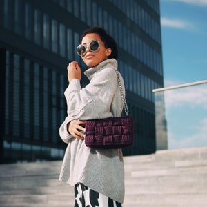 May include: A dark purple woven handbag with a silver chain strap, paired with a light gray turtleneck sweater and a black and white abstract patterned skirt. The accessory features a thick, grid-like texture and a rectangular silhouette.
