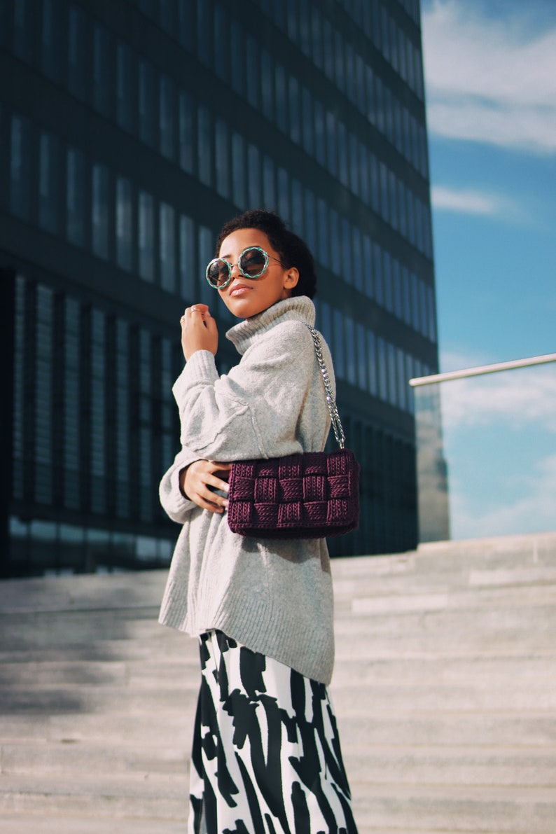 May include: A dark purple woven handbag with a silver chain strap, paired with a light gray turtleneck sweater and a black and white abstract patterned skirt. The accessory features a thick, grid-like texture and a rectangular silhouette.