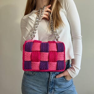 May include: A rectangular handbag featuring a woven checkerboard pattern in vibrant pink and deep purple yarn. The bag is equipped with a thick, chunky silver-toned metal chain strap that connects to the top of the bag, providing a sturdy handle.