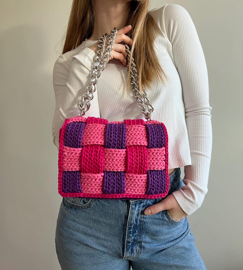 May include: A rectangular handbag featuring a woven checkerboard pattern in vibrant pink and deep purple yarn. The bag is equipped with a thick, chunky silver-toned metal chain strap that connects to the top of the bag, providing a sturdy handle.