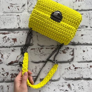 K&ouml;nnte beinhalten: Eine leuchtend neongelbe H&auml;keltasche mit einer strukturierten, geknoteten Maschenoptik. Die Tasche wird mit einem dunkelmetallischen Druckknopfverschluss gesichert und verf&uuml;gt &uuml;ber einen passenden neongelben H&auml;kelriemen mit einer dunklen Metallkette.