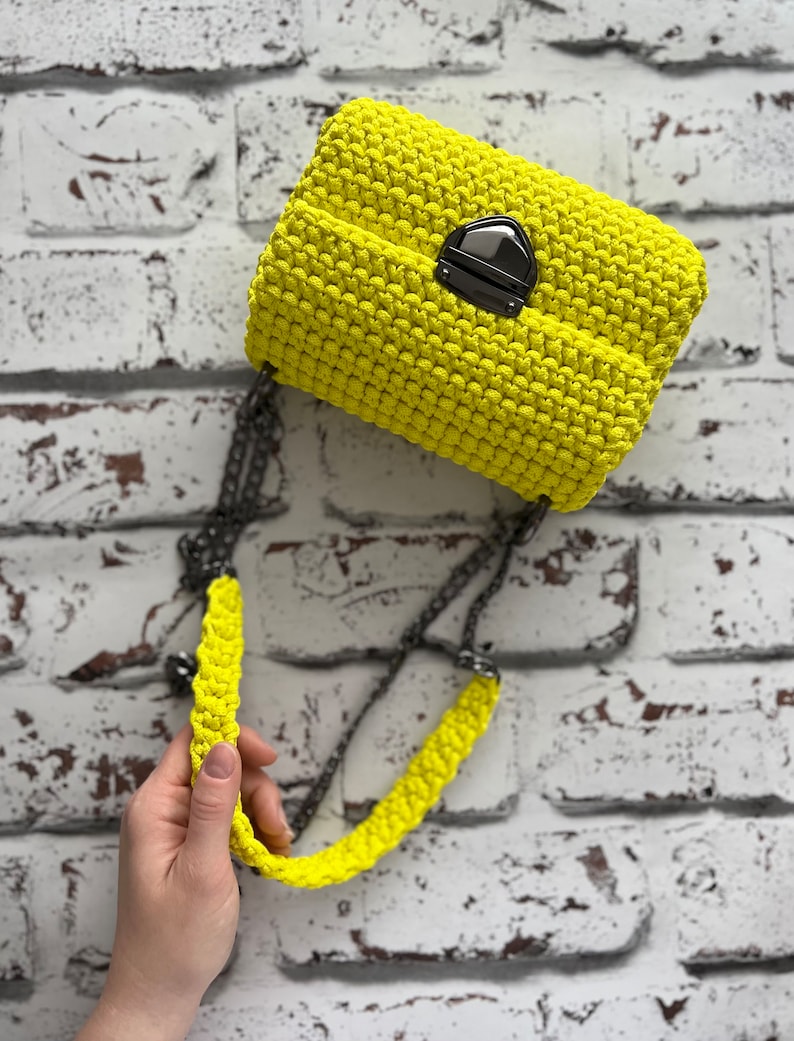 K&ouml;nnte beinhalten: Eine leuchtend neongelbe H&auml;keltasche mit einer strukturierten, geknoteten Maschenoptik. Die Tasche wird mit einem dunkelmetallischen Druckknopfverschluss gesichert und verf&uuml;gt &uuml;ber einen passenden neongelben H&auml;kelriemen mit einer dunklen Metallkette.