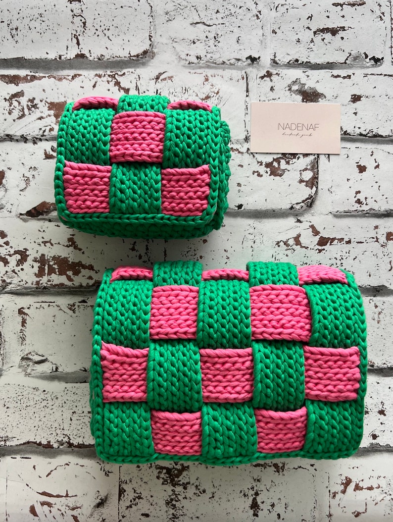 May include: Two rectangular bags crafted from thick, woven yarn in a vibrant checkerboard pattern of bright green and hot pink. The bags feature a chunky, textured knit construction with a folded flap design. A small rectangular card reads NADENAF.
