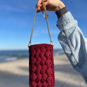 May include: A rectangular, deep burgundy crochet pouch featuring a thick, braided cord texture. The pouch is attached to a thin, gold-toned metal chain strap with lobster claw clasps, providing a functional way to carry small items securely.