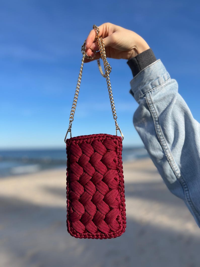 May include: A rectangular, deep burgundy crochet pouch featuring a thick, braided cord texture. The pouch is attached to a thin, gold-toned metal chain strap with lobster claw clasps, providing a functional way to carry small items securely.