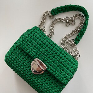 K&ouml;nnte beinhalten: Eine leuchtend gr&uuml;ne H&auml;keltasche mit einer strukturierten, gewebten Kordelkonstruktion. Die Tasche verf&uuml;gt &uuml;ber einen gl&auml;nzenden, silberfarbenen Metalldruckknopfverschluss an der Frontklappe und einen silbernen Kettenriemen, der mit gr&uuml;ner Kordel durchflochten ist.