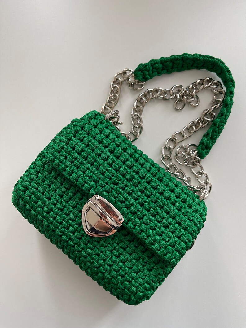 K&ouml;nnte beinhalten: Eine leuchtend gr&uuml;ne H&auml;keltasche mit einer strukturierten, gewebten Kordelkonstruktion. Die Tasche verf&uuml;gt &uuml;ber einen gl&auml;nzenden, silberfarbenen Metalldruckknopfverschluss an der Frontklappe und einen silbernen Kettenriemen, der mit gr&uuml;ner Kordel durchflochten ist.