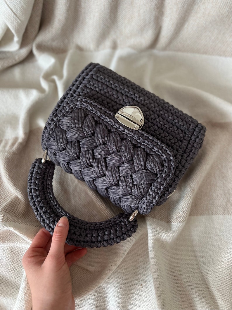 Op de afbeelding: Een donkergrijze gehaakte handtas met een gevlochten textuur op de voorflap, een afgerond handvat en een zilverkleurige metalen draaisluiting. De tas is gemaakt van dik koordmateriaal met een consistente donkergrijze kleur over het geheel.