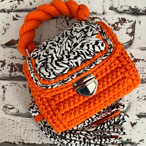 Puede incluir: Un bolso de ganchillo naranja brillante con un asa trenzada gruesa, un cierre de presi&oacute;n de metal plateado y una solapa decorativa con una trenza de patr&oacute;n blanco y negro. Un fleco largo hecho de tiras de tela blanca y negra cuelga de la base.