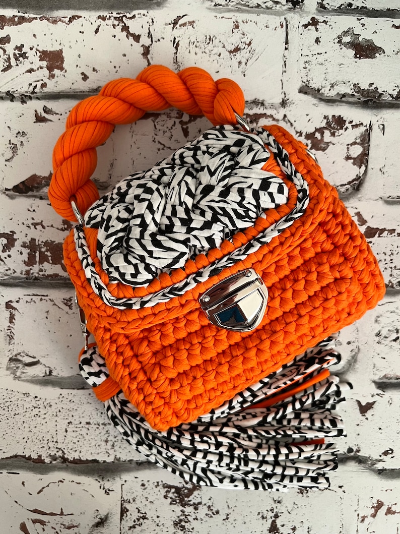 Puede incluir: Un bolso de ganchillo naranja brillante con un asa trenzada gruesa, un cierre de presi&oacute;n de metal plateado y una solapa decorativa con una trenza de patr&oacute;n blanco y negro. Un fleco largo hecho de tiras de tela blanca y negra cuelga de la base.