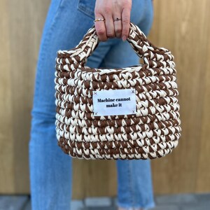 May include: A hand-crocheted handbag featuring a two-tone pattern of alternating dark brown and cream yarn. The bag has a rectangular shape with integrated handles and a white fabric label stitched to the front with the text Machine cannot make it.
