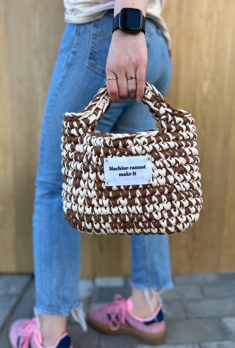 May include: A hand-crocheted handbag featuring a two-tone pattern of alternating dark brown and cream yarn. The bag has a rectangular shape with integrated handles and a white fabric label stitched to the front with the text Machine cannot make it.