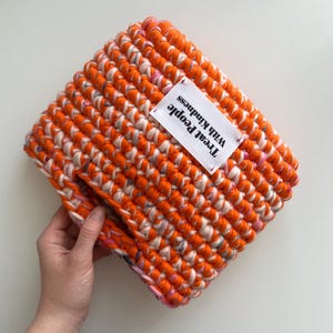 May include: A rectangular crochet pouch made from thick, vibrant orange and variegated pink and white yarn. A white rectangular fabric label is stitched onto the front, featuring the black printed text, Treat People With Kindness.