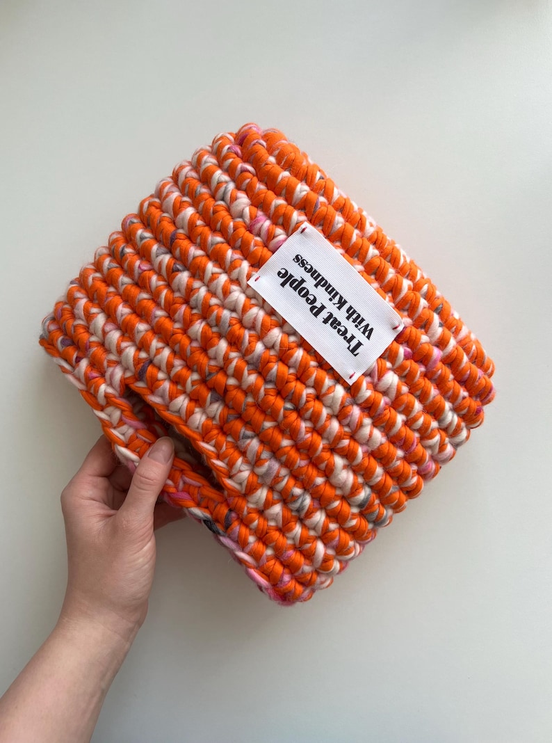 May include: A rectangular crochet pouch made from thick, vibrant orange and variegated pink and white yarn. A white rectangular fabric label is stitched onto the front, featuring the black printed text, Treat People With Kindness.