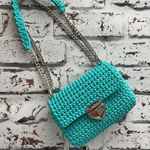 K&ouml;nnte beinhalten: Eine t&uuml;rkisfarbene H&auml;keltasche mit strukturiertem Maschenmuster, einem silberfarbenen Metalldrehverschluss und einem zweigeteilten Trageriemen aus einem passenden t&uuml;rkisfarbenen H&auml;kel-Schulterpolster und einer polierten silberfarbenen Metallkette.