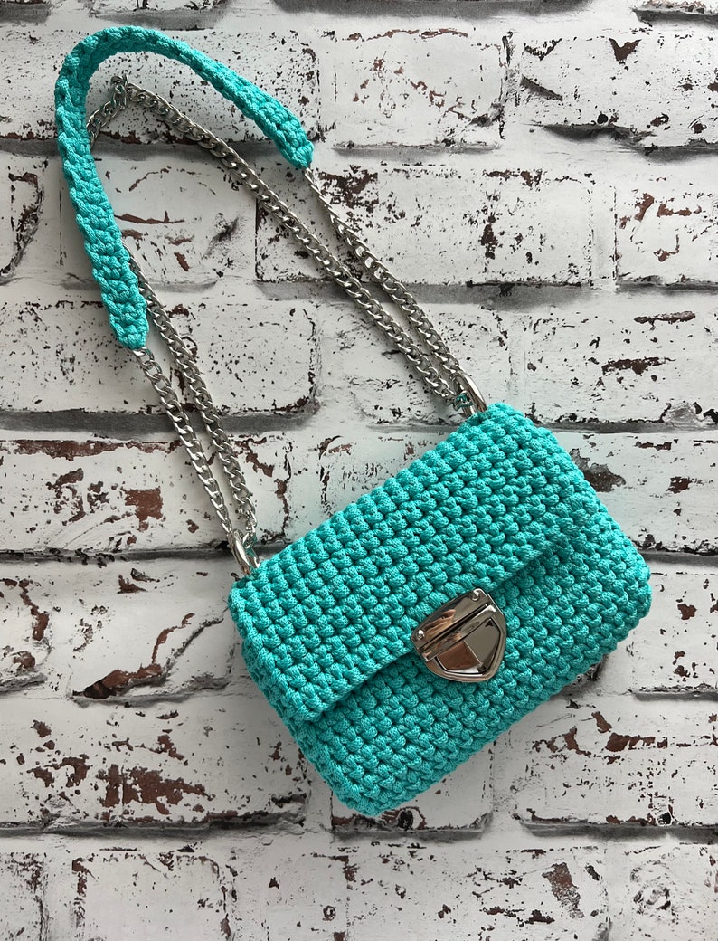 K&ouml;nnte beinhalten: Eine t&uuml;rkisfarbene H&auml;keltasche mit strukturiertem Maschenmuster, einem silberfarbenen Metalldrehverschluss und einem zweigeteilten Trageriemen aus einem passenden t&uuml;rkisfarbenen H&auml;kel-Schulterpolster und einer polierten silberfarbenen Metallkette.