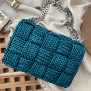 May include: A teal handbag featuring a thick, woven, chunky knit texture. The bag has a rectangular shape and includes a silver-toned metal chain strap attached to the top. The material appears to be a soft, cord-like yarn woven in a grid pattern.