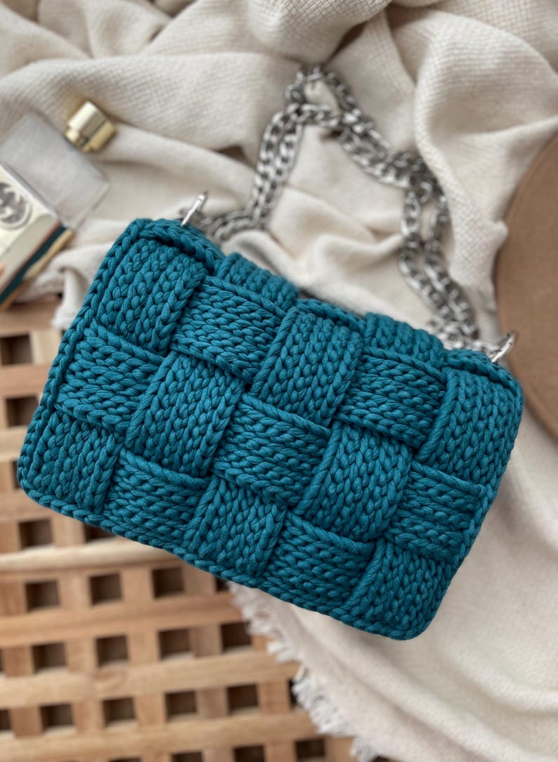 May include: A teal handbag featuring a thick, woven, chunky knit texture. The bag has a rectangular shape and includes a silver-toned metal chain strap attached to the top. The material appears to be a soft, cord-like yarn woven in a grid pattern.