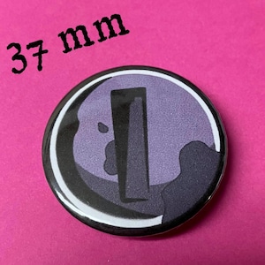 May include: A round button badge with a black and white design. The design features a purple silhouette of a person with a large, rectangular shape in the center. The text "37 mm" is printed above the badge.