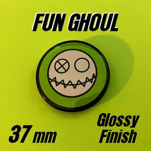 May include: A circular pin button with a glossy finish measuring 37 mm in diameter. The design features a white face with a stitched mouth, one eye as an X, and one eye as a circle, set against a bright green background with the text FUN GHOUL.