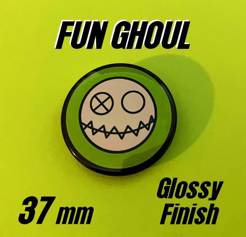 May include: A circular pin button with a glossy finish measuring 37 mm in diameter. The design features a white face with a stitched mouth, one eye as an X, and one eye as a circle, set against a bright green background with the text FUN GHOUL.
