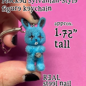 May include: A bright blue flocked animal figure keychain with black ear details and dark eye markings. The figure features a small silver-colored metal nail embedded in its chest and measures approximately 1.75 inches tall. Text reads Flocked Sylvanian-Style figure keychain and REAL steel nail.