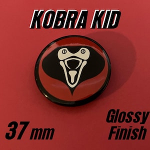 May include: A circular pin with a glossy finish featuring a stylized black and cream cobra head graphic on a red background. The text KOBRA KID is printed above the pin, and the text 37 mm GLOSSY FINISH is printed below the pin.