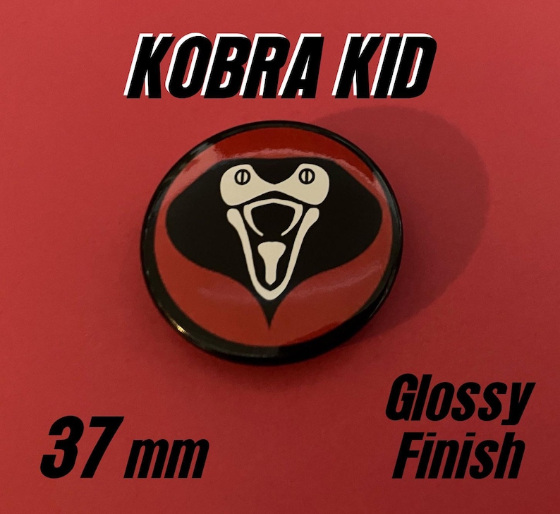 May include: A circular pin with a glossy finish featuring a stylized black and cream cobra head graphic on a red background. The text KOBRA KID is printed above the pin, and the text 37 mm GLOSSY FINISH is printed below the pin.