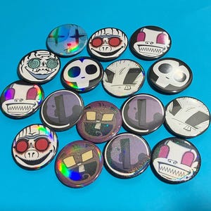 May include: A collection of circular pin-back buttons featuring various graphic illustrations, including stylized skulls, cartoon faces with red eyes, and abstract shapes. Several buttons have a holographic or glitter finish.