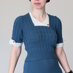 May include: A blue knitted outfit comprising a beret, a top with short sleeves, and a skirt. The top has a white collar and cuffs with red button details. The ensemble has a textured pattern and is made of a soft knit material.