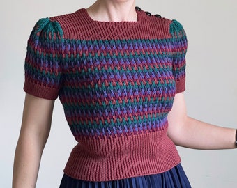 May include: A red, green, purple and black short-sleeved sweater with a geometric pattern. The sweater has a round neckline and a fitted silhouette.
