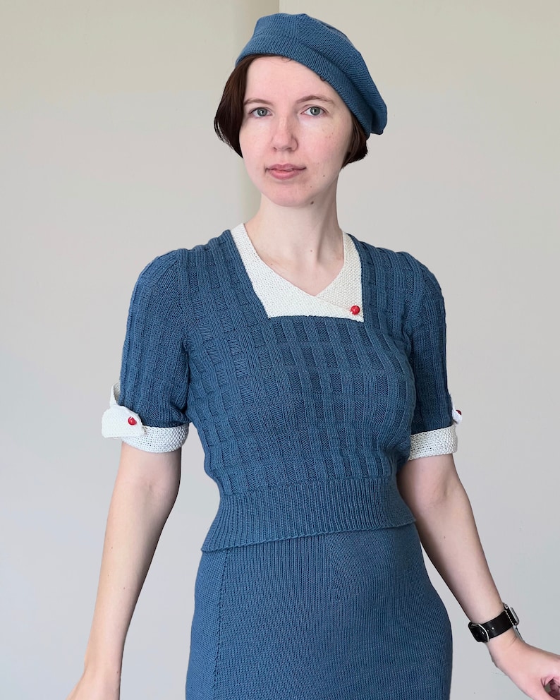 May include: A blue knitted outfit comprising a beret, a top with short sleeves, and a skirt. The top has a white collar and cuffs with red button details. The ensemble has a textured pattern and is made of a soft knit material.