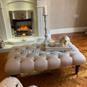 May include: A beige tufted footstool with wooden legs sits in front of a fireplace. A white tray with a candle and a decorative skull sits on top of the footstool. The text "We Make Footstools" is visible on the footstool.