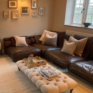 May include: A large brown leather sectional sofa with a cream-coloured ottoman in front of it. The ottoman has a tufted top and wooden legs. The sofa has several cushions on it, including a cream-coloured cushion with a brown and cream patterned cushion on top of it. The sofa is in a living room with a window and a wall with framed pictures.