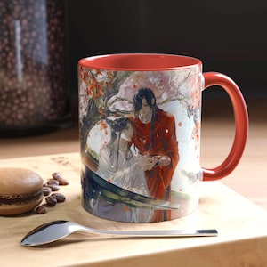 May include: A ceramic coffee mug with a red interior and handle. The exterior features a detailed illustration of two figures in traditional attire under a flowering tree. The mug is set on a wooden surface with a spoon and a macaron.