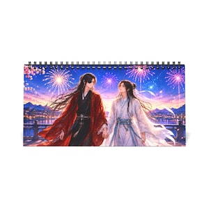 Puede incluir: Un calendario de escritorio horizontal con encuadernación en espiral que muestra una ilustración de dos figuras con túnicas tradicionales, una roja y otra blanca, tomadas de la mano bajo un cielo nocturno lleno de fuegos artificiales morados y dorados.
