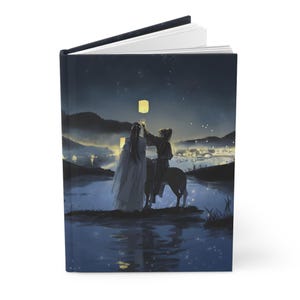 Puede incluir: Diario de tapa dura con una ilustración nocturna. Dos figuras sueltan linternas sobre el agua, con un pueblo a lo lejos. La portada muestra un cielo azul oscuro, un lago reflectante y una ciudad iluminada.