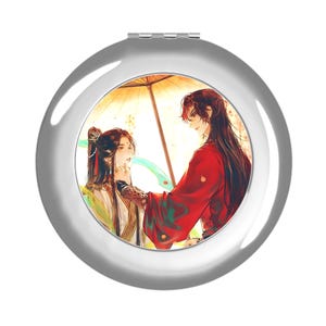 May include: A round, silver-toned compact mirror featuring a circular illustration of two figures under a golden umbrella. One figure wears a red robe, while the other wears white and gold attire. The artwork displays long dark hair and flowing ribbons.