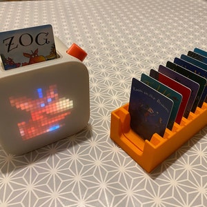 May include: A white and orange children's book reader with a pixelated image of a dragon on the front. The reader has a slot for a book card. A separate orange card holder with 8 book cards, including one with the title "Room on the Broom", is shown.