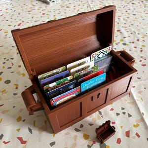 May include: A brown wooden treasure chest with a hinged lid is open and filled with colourful book cards. The cards have titles like "Mog", "Charlie Cook's Favourite", and "The Christmasaurus".