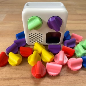 May include: A white box with two knobs, one green and one purple, and a speaker grille. Surrounding the box are 16 colorful knobs in various colors, including red, yellow, blue, green, purple, and pink.