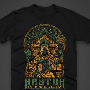 Op de afbeelding: Zwart T-shirt met een gedetailleerde afbeelding van een geklede figuur met een kroon, zittend voor een kasteel. Het ontwerp bevat de tekst "HASTUR THE KING IN YELLOW" en andere zinnen. Het kunstwerk is in tinten groen, geel en oranje.