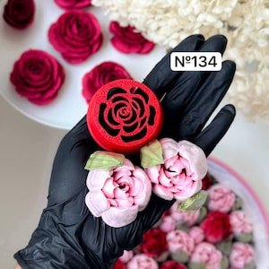 以下が含まれることがあります： バタークリームの花を作るための赤いプラスチック製のバラ型のモールド。モールドは黒い手袋をした手で持たれています。背景にはピンクと赤のバタークリームのバラがいくつかあります。