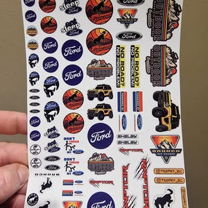 May include: A sheet of Ford-themed stickers featuring various designs. The stickers include the Ford logo, Bronco graphics, and phrases like "Outdoor Expedition" and "No Road? No Problem!" in a variety of colors.